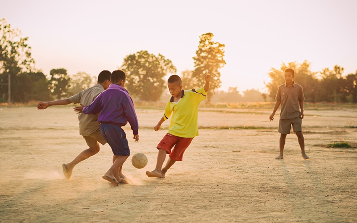 Young players kicking a ball on an open dirt field at sunset